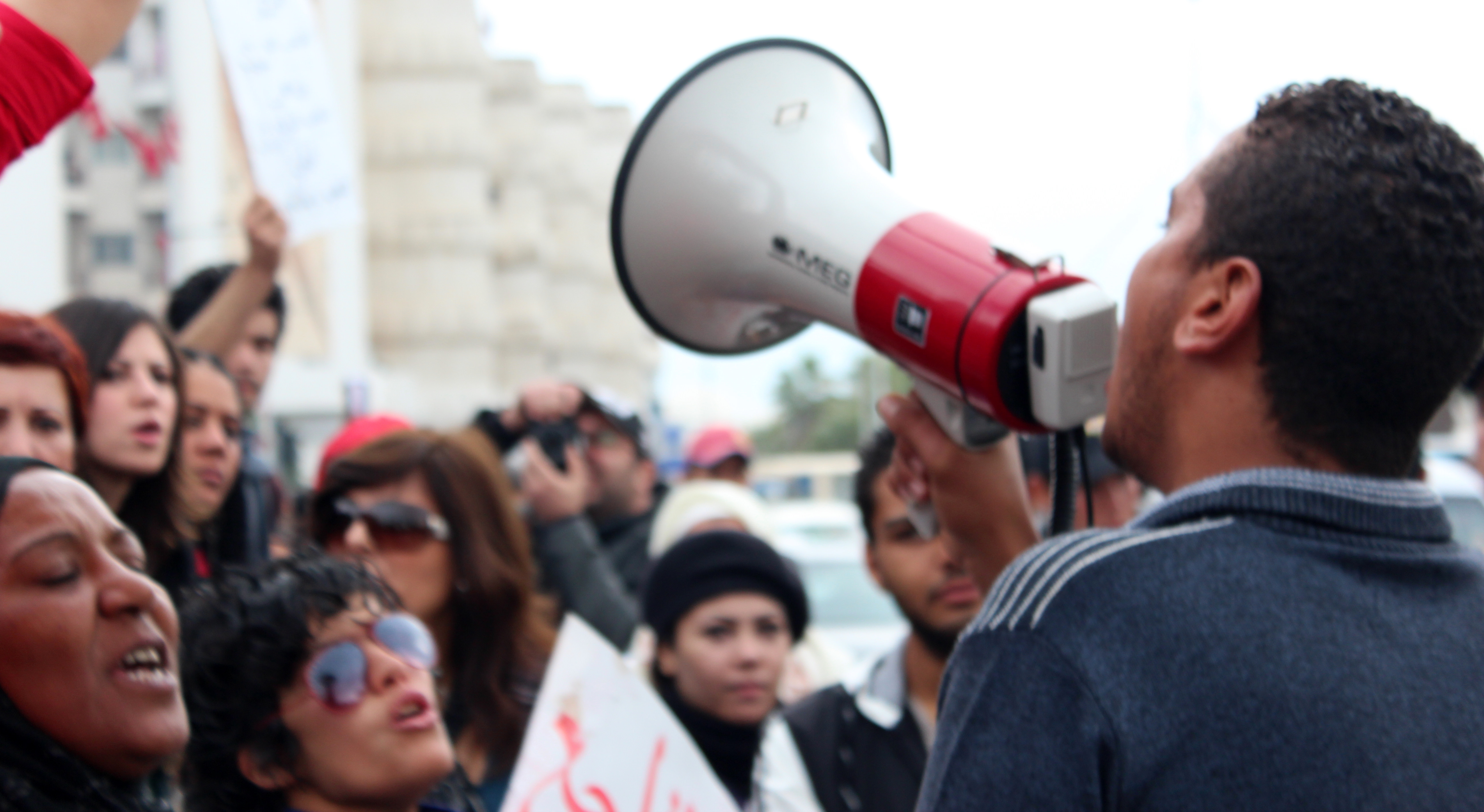 Man faces away from camera, holding a white and red bullhorn. People stand in front of him, seeming to speak along with him.